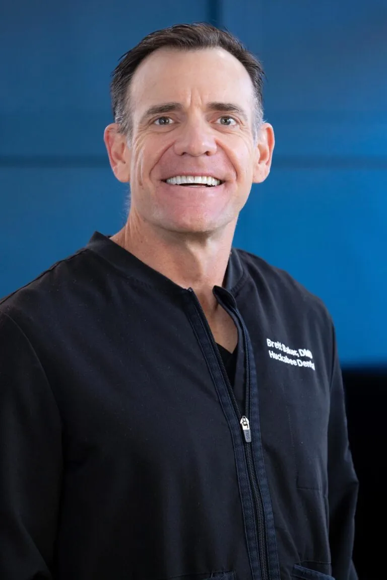 Headshot of Dr. Brett Baker wearing a black jacket against a solid blue background. The image emphasizes their role in dental and maxillofacial care, presenting a polished and professional appearance.
