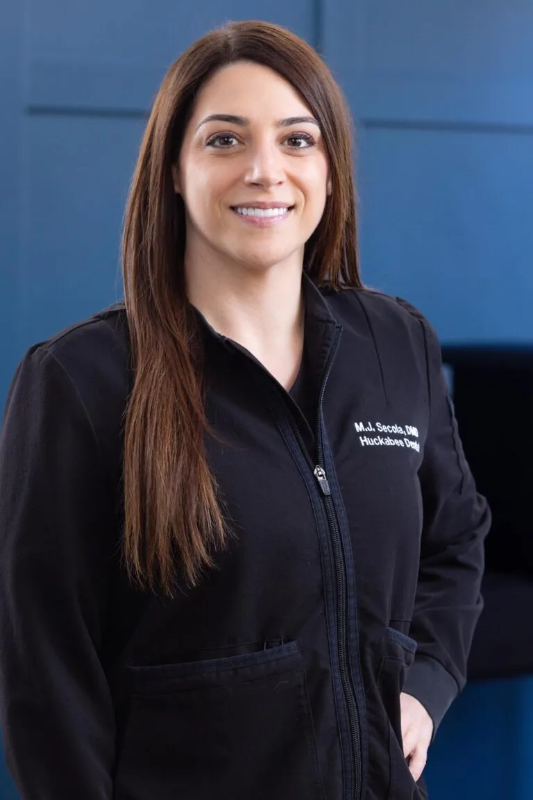 Headshot of Dr. Mj Secola wearing a black jacket with embroidered on the left side stands against a solid blue background. The focusing attention on the name and branding elements of the image.