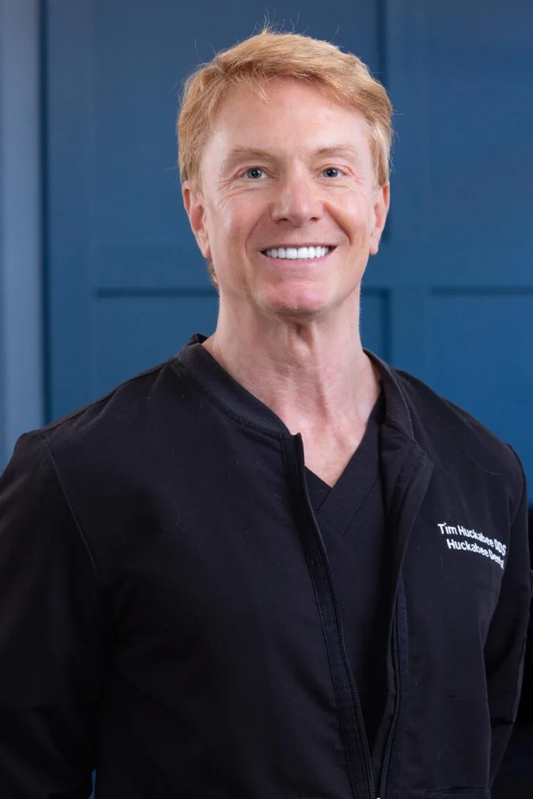 Headshot Dr. Tim Huckabee wearing a black jacket with embroidered on the left side stands against a blue panel background. The image emphasizes their role as a medical doctor, highlighting their name and affiliation.