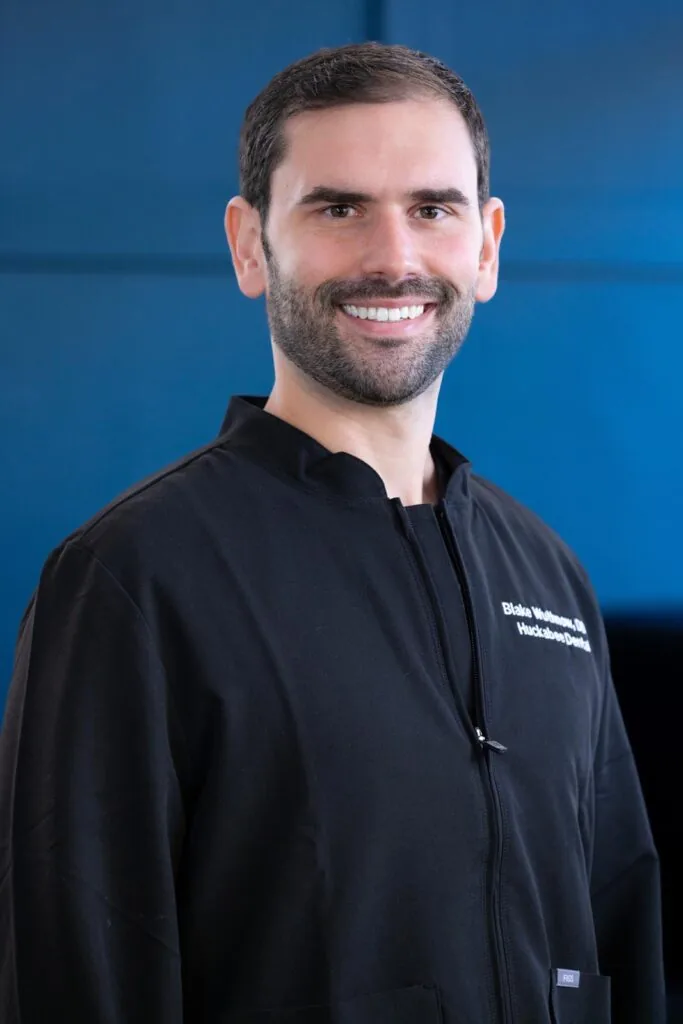 Headshot of Dr. Blake Wuthnow wearing a black jacket with embroidered on the left side stands against a solid blue background. The image emphasizes a personalized title associated with the individual's identity, presenting a sharp and formal look.