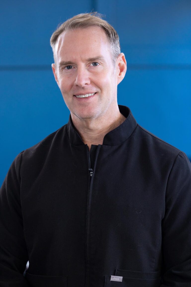Headshot of Dr. Tom Draper wearing a black zip-up jacket stands against a solid blue background, with a small tag visible near the bottom. The image emphasizes professional attire and a clean, minimal composition.