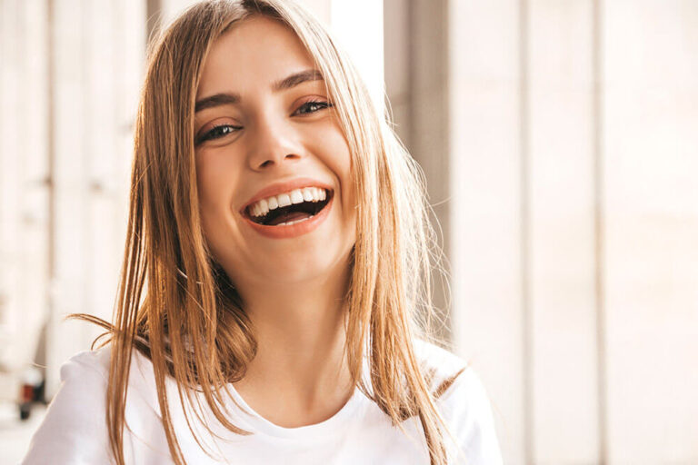 A person wearing a white shirt sits indoors, with long, straight hair framing their face. The background is blurred, showing windows or walls, keeping focus on the subject.