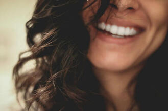 Close-up of a wide smile showing white teeth and faint dimples, partially framed by curly dark hair. The softly blurred background emphasizes the joyful expression.