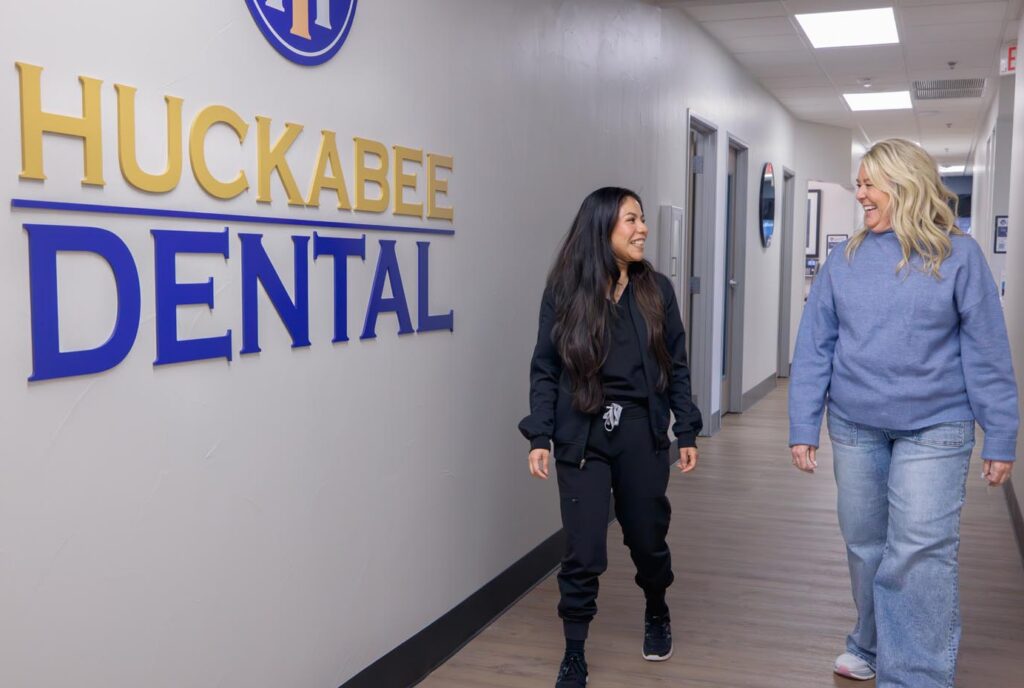 Two dental staff walking through the Huckabee Dental hallway, smiling and talking near the clinic logo.